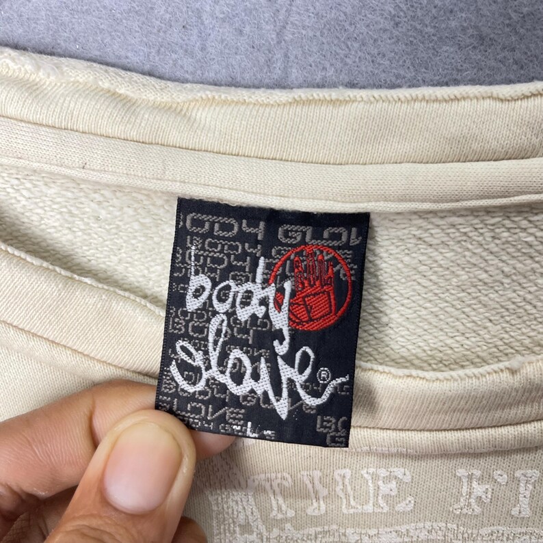 Puede incluir: Una sudadera de color crema con una etiqueta de tela rectangular negra cosida en el interior del cuello. La etiqueta muestra el texto blanco "body glove" en una fuente cursiva estilizada, junto a un logotipo circular rojo.