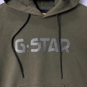 Puede incluir: Sudadera con capucha verde oliva con el texto "G-STAR" en gris oscuro texturizado en el pecho. La sudadera tiene una capucha con cord&oacute;n y un bolsillo de canguro. La tela parece ser un material suave y de peso medio.