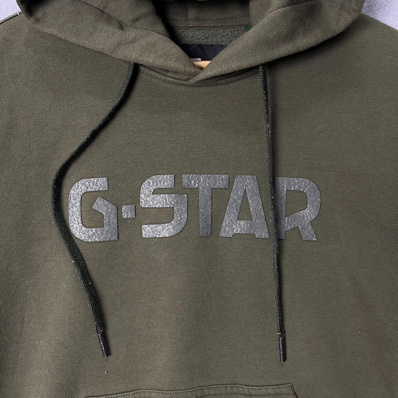 Puede incluir: Sudadera con capucha verde oliva con el texto "G-STAR" en gris oscuro texturizado en el pecho. La sudadera tiene una capucha con cord&oacute;n y un bolsillo de canguro. La tela parece ser un material suave y de peso medio.
