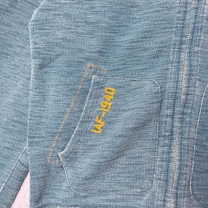 Peut inclure: Gros plan sur une veste bleu clair avec un tissage textur&eacute;. La veste comporte une poche avec le texte brod&eacute; "AF-1940" en jaune. Une fermeture &eacute;clair parcourt l'avant de la veste.