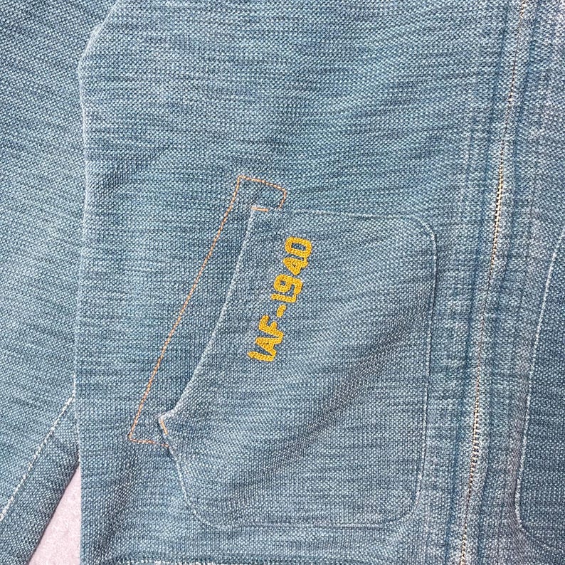 Peut inclure: Gros plan sur une veste bleu clair avec un tissage textur&eacute;. La veste comporte une poche avec le texte brod&eacute; "AF-1940" en jaune. Une fermeture &eacute;clair parcourt l'avant de la veste.