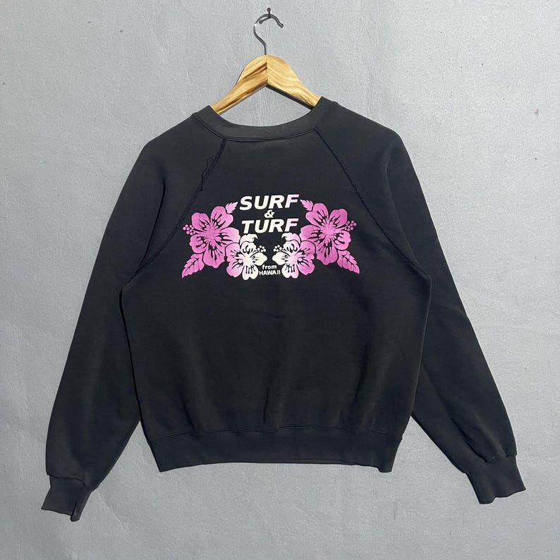 Op de afbeelding: Zwart sweatshirt met een ronde hals en lange mouwen. De voorkant heeft een bloemenontwerp in roze en wit, met de woorden "SURF & TURF" erboven en "From HAWAII" onder de bloemen. Het sweatshirt hangt aan een houten hanger.