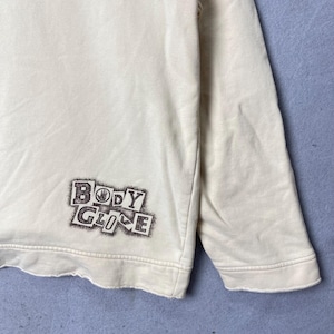 Puede incluir: Una sudadera de manga larga color crema con un estampado gr&aacute;fico marr&oacute;n oscuro en la parte inferior izquierda. El gr&aacute;fico muestra el texto "BODY GLOVE" en una fuente de estilo bloque desgastado con un icono de mano en la letra O.
