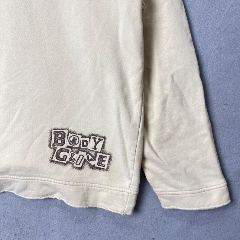 Puede incluir: Una sudadera de manga larga color crema con un estampado gr&aacute;fico marr&oacute;n oscuro en la parte inferior izquierda. El gr&aacute;fico muestra el texto "BODY GLOVE" en una fuente de estilo bloque desgastado con un icono de mano en la letra O.