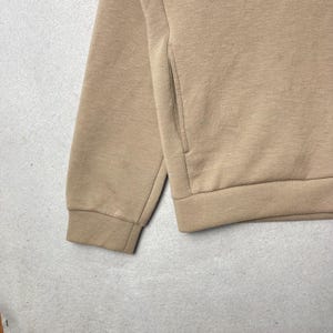 May include: A tan-colored sweatshirt with a pocket and ribbed cuffs. The image shows a close-up of the sleeve and part of the body of the garment. The fabric appears to be a soft, knit material.
