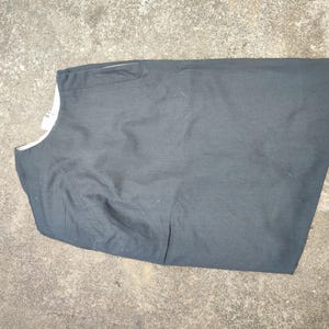 May include: A dark gray vest with a white lining visible at the neckline. The vest is lying flat on a concrete surface. The fabric appears to be a woven material.