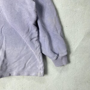 May include: A close-up of a lavender-colored long-sleeved shirt. The shirt has a ribbed cuff on the sleeve and a faded appearance. The fabric appears to be a soft, comfortable material.
