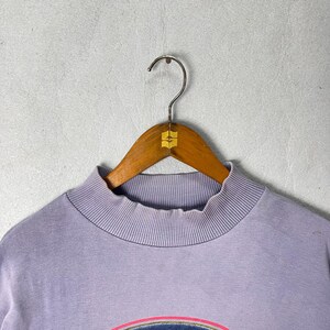 May include: A light purple sweatshirt with a ribbed mock neck and a graphic design at the bottom. The sweatshirt is hanging on a wooden hanger with a metal hook.