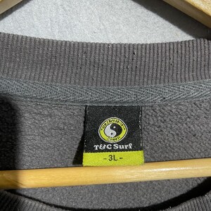 Puede incluir: Primer plano de una etiqueta de sudadera gris con el texto "T&C Surf" y un s&iacute;mbolo de yin-yang. La etiqueta es verde y negra con la talla "-3L-" impresa debajo del nombre de la marca. La sudadera es de color gris apagado.