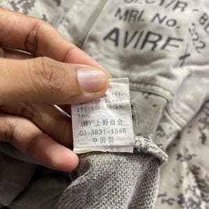 Puede incluir: Primer plano de una chaqueta con cremallera gris claro con la marca "AVIREX" impresa. La chaqueta tiene un patr&oacute;n de camuflaje y una etiqueta con texto japon&eacute;s y el texto "03-3831-1546".