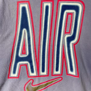 May include: Close-up of a lilac-colored t-shirt featuring the word "AIR" in large, bold letters. The letters are outlined in red and white, with a dark blue interior. A gold and black Nike swoosh logo is at the bottom.