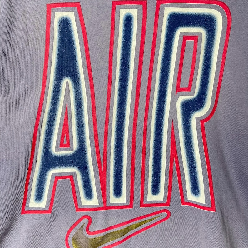 May include: Close-up of a lilac-colored t-shirt featuring the word "AIR" in large, bold letters. The letters are outlined in red and white, with a dark blue interior. A gold and black Nike swoosh logo is at the bottom.
