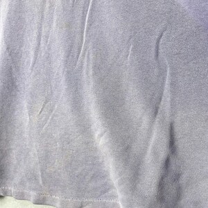 May include: Close-up of a light purple t-shirt. The fabric is slightly wrinkled and has some discoloration. The bottom hem is visible. The shirt appears to be made of a soft, knit material.