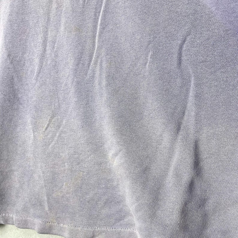 May include: Close-up of a light purple t-shirt. The fabric is slightly wrinkled and has some discoloration. The bottom hem is visible. The shirt appears to be made of a soft, knit material.