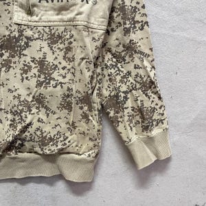 Puede incluir: Sudadera de camuflaje en tonos beige y marr&oacute;n con cuello con cremallera. La sudadera tiene pu&ntilde;os y dobladillo de canal&eacute;. La palabra "AVIREX" est&aacute; impresa en la parte superior del pecho.