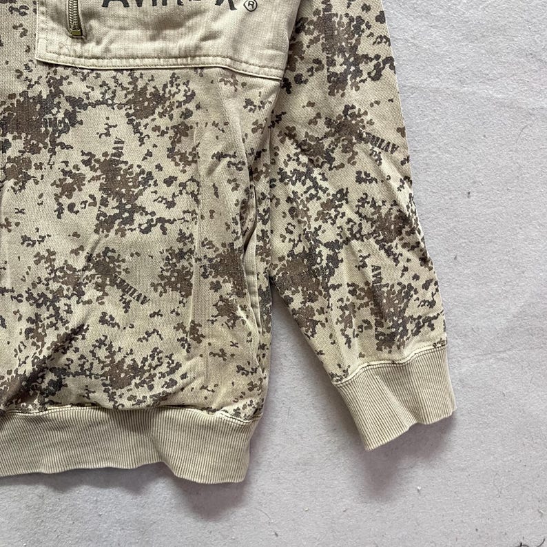 Puede incluir: Sudadera de camuflaje en tonos beige y marr&oacute;n con cuello con cremallera. La sudadera tiene pu&ntilde;os y dobladillo de canal&eacute;. La palabra "AVIREX" est&aacute; impresa en la parte superior del pecho.