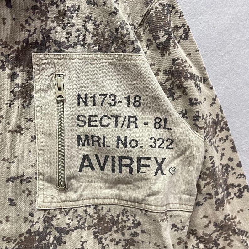 Puede incluir: Primer plano de una chaqueta con estampado de camuflaje en beige y marr&oacute;n. Un bolsillo rectangular con cremallera en la parte delantera, con el texto "N173-18 SECT/R - 8L MRI. No. 322 AVIREX&reg;" en negro.