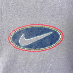 May include: A light purple t-shirt featuring a large, oval-shaped logo. The logo has a blue center with a white Nike swoosh, outlined by a gold and pink border. The shirt's fabric has a slightly textured appearance.