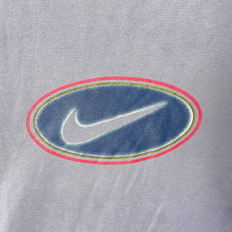 May include: A light purple t-shirt featuring a large, oval-shaped logo. The logo has a blue center with a white Nike swoosh, outlined by a gold and pink border. The shirt's fabric has a slightly textured appearance.
