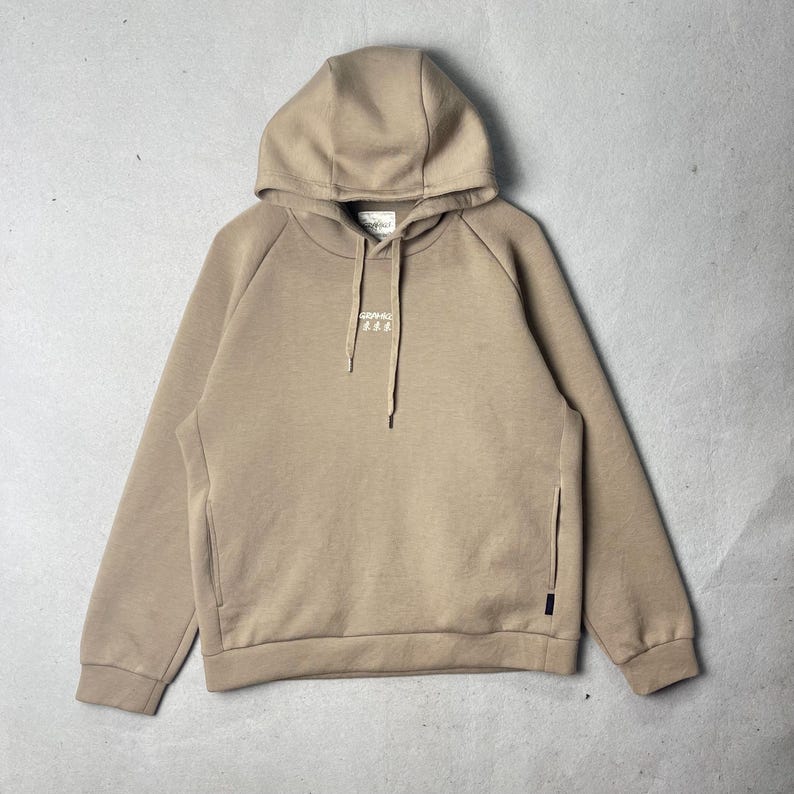 May include: A tan hoodie with a drawstring hood and a kangaroo pocket. The hoodie has a small white logo on the chest. The sleeves are long and the material appears to be a soft fabric. The hoodie is laid flat on a gray surface.