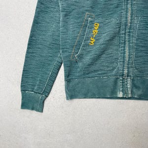 Peut inclure: Sweat &agrave; capuche zipp&eacute; bleu sarcelle avec une poche avant et le texte "IAF-1940" en jaune. Le sweat &agrave; capuche a un aspect vieilli avec un poignet et un ourlet c&ocirc;tel&eacute;s. La fermeture &eacute;clair est visible au centre.
