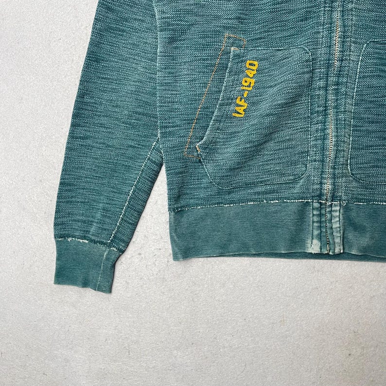 Peut inclure: Sweat &agrave; capuche zipp&eacute; bleu sarcelle avec une poche avant et le texte "IAF-1940" en jaune. Le sweat &agrave; capuche a un aspect vieilli avec un poignet et un ourlet c&ocirc;tel&eacute;s. La fermeture &eacute;clair est visible au centre.