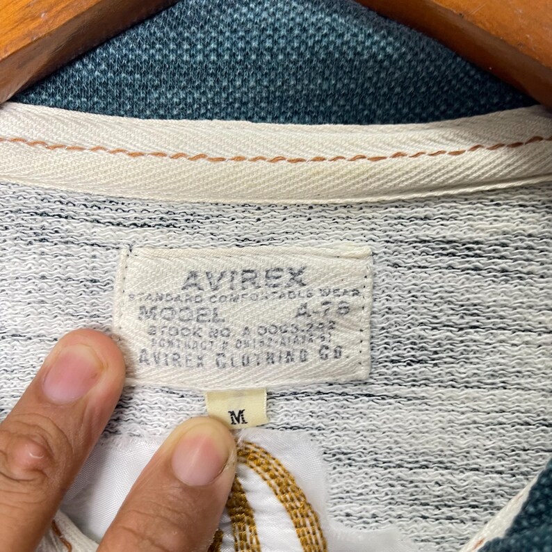 Peut inclure: Gros plan sur une &eacute;tiquette de chemise Avirex grise et blanche. L'&eacute;tiquette indique "AVIREX STANDARD COMFORTABLE WEAR MODEL A-78 STOCK NO. A 0043-292 AVIREX CLOTHING CO." Une &eacute;tiquette de taille "M" est visible. La chemise a un col vert.