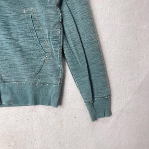 Peut inclure: Gros plan d'un sweat &agrave; capuche de couleur sarcelle avec une poche et des poignets c&ocirc;tel&eacute;s. Le tissu a un aspect textur&eacute; avec des d&eacute;tails de couture visibles. La conception comprend une capuche &agrave; cordon et une poche kangourou.