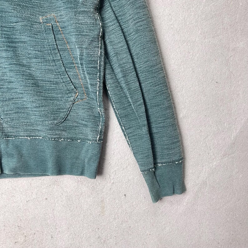 Peut inclure: Gros plan d'un sweat &agrave; capuche de couleur sarcelle avec une poche et des poignets c&ocirc;tel&eacute;s. Le tissu a un aspect textur&eacute; avec des d&eacute;tails de couture visibles. La conception comprend une capuche &agrave; cordon et une poche kangourou.
