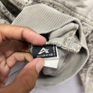 Puede incluir: Primer plano de una sudadera beige y gris con estampado de camuflaje. La etiqueta de la marca Avirex es visible, con fondo negro, logotipo y texto blancos. La tela de la sudadera parece suave y texturizada.