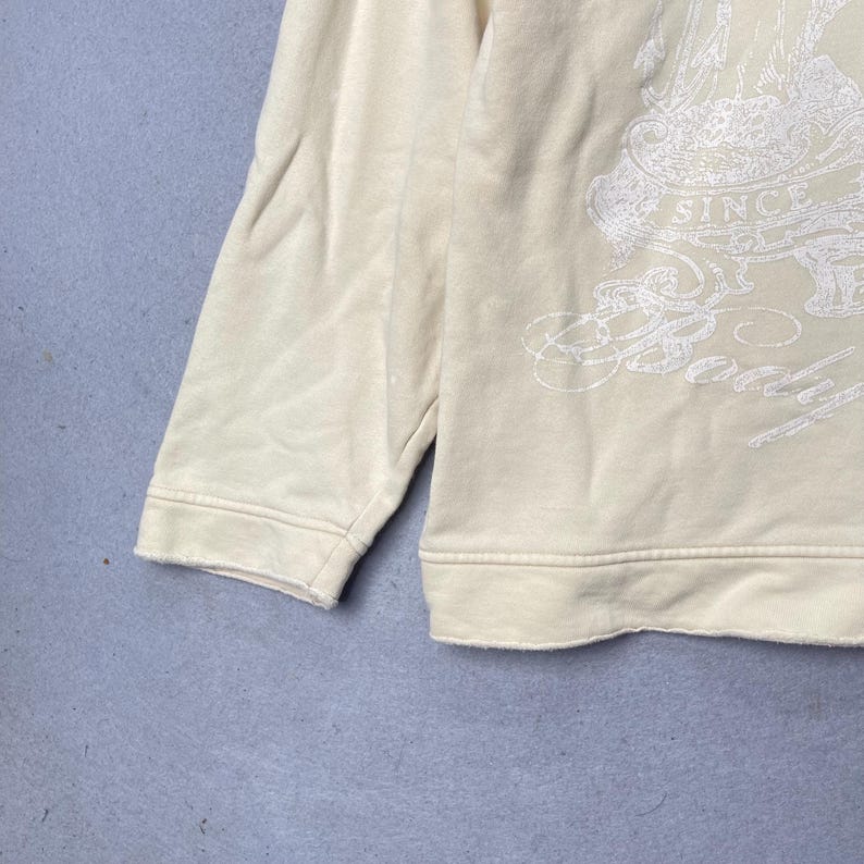 Puede incluir: Una sudadera de manga larga de color crema con un estampado gr&aacute;fico blanco en la parte delantera. El dise&ntilde;o incluye el texto "SINCE" y "Body" en una fuente cursiva estilizada. La prenda tiene cuello redondo, pu&ntilde;os acanalados y un dobladillo acanalado.