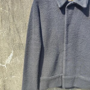 May include: A dark grey jacket with a textured finish, a collar, and long sleeves. The jacket is buttoned and appears to be made of a thick, warm material. The jacket is hanging on a wooden hanger.