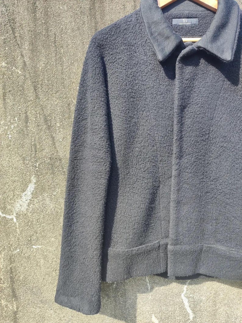 May include: A dark grey jacket with a textured finish, a collar, and long sleeves. The jacket is buttoned and appears to be made of a thick, warm material. The jacket is hanging on a wooden hanger.