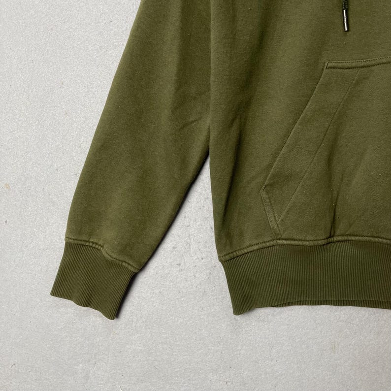 Puede incluir: Sudadera con capucha verde oliva con bolsillo de canguro y pu&ntilde;os y dobladillo acanalados. La sudadera tiene una capucha con cord&oacute;n y un dise&ntilde;o cl&aacute;sico. La tela parece ser un material suave y de peso medio.