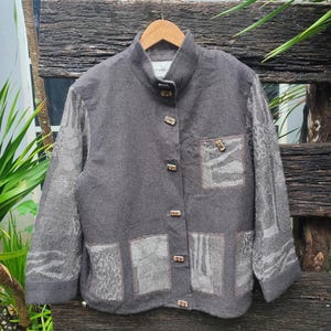 Peut inclure: Veste grise à col montant avec des boutons en bois. La veste présente un tissu à motifs sur les manches et des patchs sur le devant. Elle est suspendue à un cintre en bois sur un fond en bois rustique.