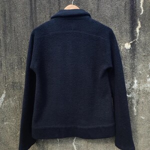 May include: A dark navy blue jacket with a textured, fleece-like finish. The jacket features a collar, long sleeves, and a simple design. It is suspended on a wooden hanger against a neutral backdrop.