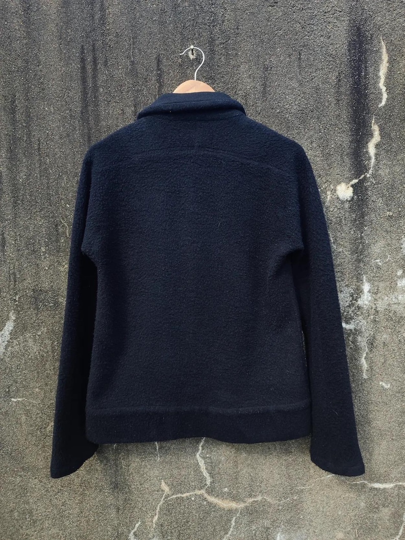 May include: A dark navy blue jacket with a textured, fleece-like finish. The jacket features a collar, long sleeves, and a simple design. It is suspended on a wooden hanger against a neutral backdrop.