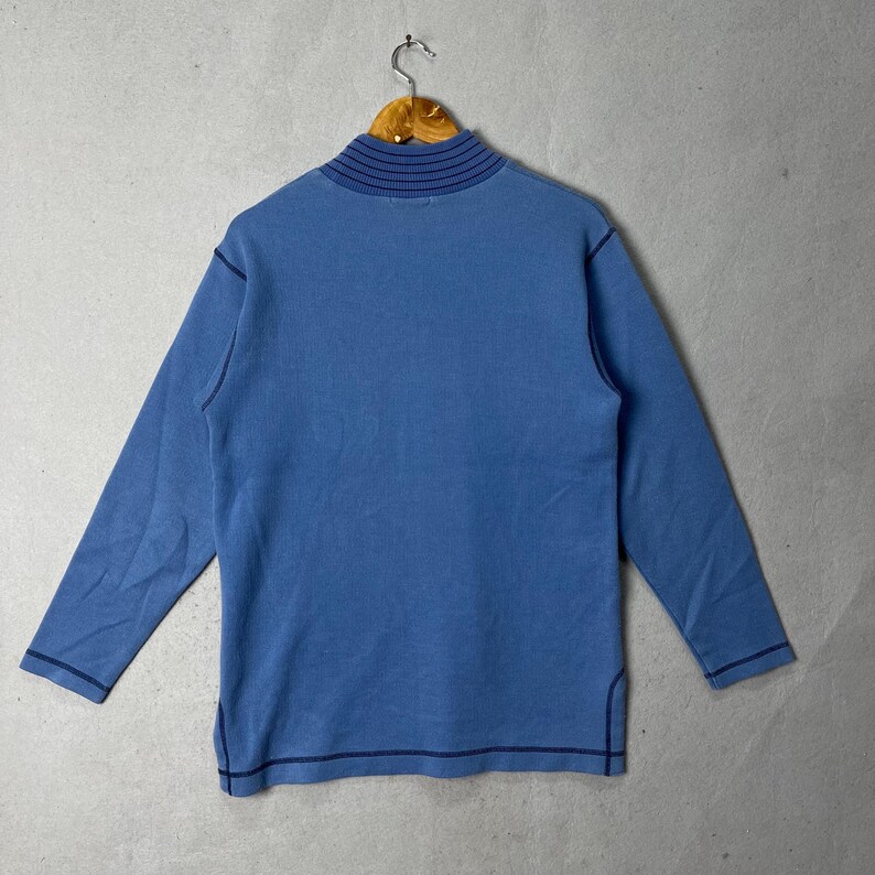 Puede incluir: Una camisa azul de manga larga con cuello alto. La camisa tiene costuras azul oscuro a lo largo de las costuras y el cuello. La camisa est&aacute; colgada en una percha de madera.