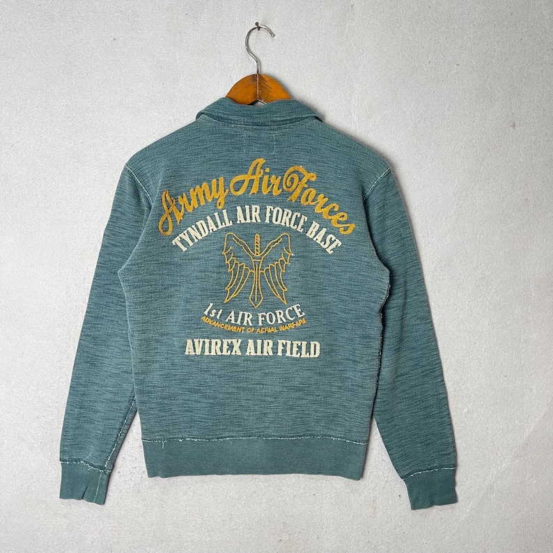 Peut inclure: Veste vintage de couleur sarcelle avec un col et des manches longues. Le dos pr&eacute;sente des lettres dor&eacute;es : "Army Air Forces, Tyndall Air Force Base, 1st Air Force, Avirex Air Field" et un embl&egrave;me ail&eacute;. La veste est suspendue &agrave; un cintre en bois.