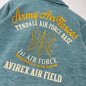 Peut inclure: Gros plan sur une veste bleu sarcelle avec des broderies. On peut lire "Army Air Forces", "Tyndall Air Force Base", "1st Air Force" et "Avirex Air Field". Le motif est une &eacute;p&eacute;e ail&eacute;e.