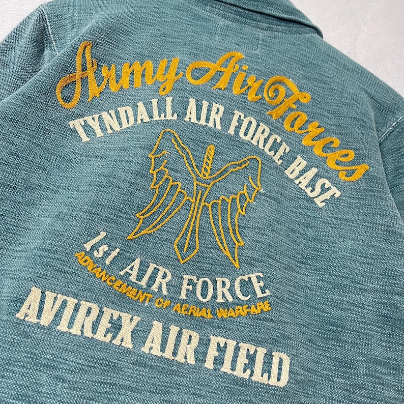 Peut inclure: Gros plan sur une veste bleu sarcelle avec des broderies. On peut lire "Army Air Forces", "Tyndall Air Force Base", "1st Air Force" et "Avirex Air Field". Le motif est une &eacute;p&eacute;e ail&eacute;e.