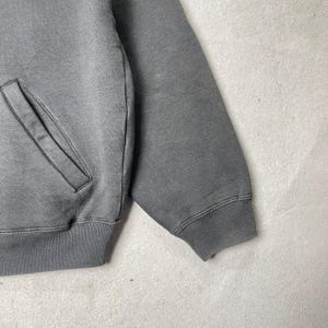 Puede incluir: Primer plano de una sudadera gris con un bolsillo y pu&ntilde;os acanalados. La prenda es de color gris liso y apagado, con un aspecto ligeramente descolorido. La manga es visible, mostrando el detalle del pu&ntilde;o acanalado.
