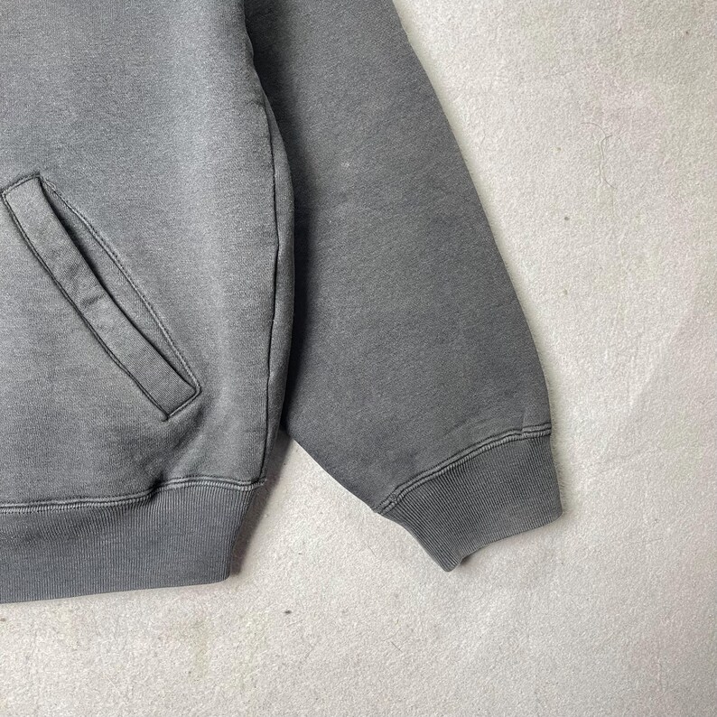 Puede incluir: Primer plano de una sudadera gris con un bolsillo y pu&ntilde;os acanalados. La prenda es de color gris liso y apagado, con un aspecto ligeramente descolorido. La manga es visible, mostrando el detalle del pu&ntilde;o acanalado.