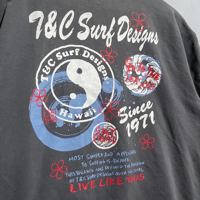 Puede incluir: Camiseta gris oscuro con un dise&ntilde;o gr&aacute;fico que presenta el texto "7&C Surf Designs" y las palabras "Hawaii" y "Since 1971". El dise&ntilde;o incluye un s&iacute;mbolo de yin-yang y acentos florales. La camiseta tambi&eacute;n tiene el texto "Live Like This".
