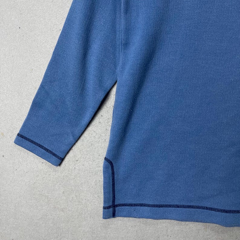Puede incluir: Una camisa azul de manga tres cuartos. La camisa tiene cuello redondo y est&aacute; hecha de una tela suave y texturizada. Las costuras est&aacute;n reforzadas con pespuntes azul oscuro.