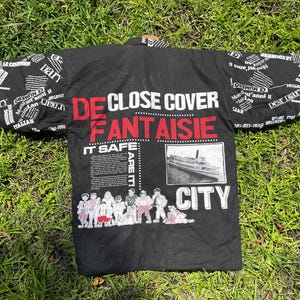 May include: Black jacket with white text on the sleeves and back. The back features the words "DE CLOSE COVER FANTAISIE" in red and white, along with a vintage ship image and the word "CITY". The sleeves have a collage of text.