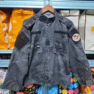 Peut inclure: Veste de camouflage gris foncé avec plusieurs poches et écussons. La veste est suspendue à un cintre en bois. La veste présente un motif unique et un design d'inspiration militaire.