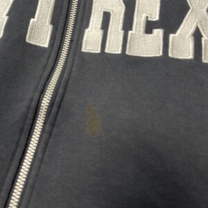 Puede incluir: Primer plano de una sudadera con capucha azul marino con cremallera y letras bordadas en blanco. La cremallera es plateada. Una mancha marr&oacute;n es visible en la parte delantera de la sudadera.