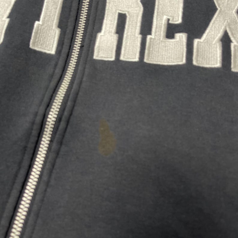 Puede incluir: Primer plano de una sudadera con capucha azul marino con cremallera y letras bordadas en blanco. La cremallera es plateada. Una mancha marr&oacute;n es visible en la parte delantera de la sudadera.