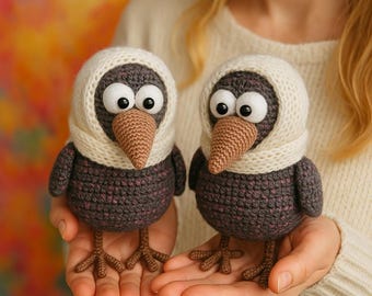 May include: Two handmade crochet birds with large googly eyes, brown beaks, and cream-colored scarves. The birds are primarily dark gray with hints of pink and brown. They have detailed crochet feet and are held in someone's hands.
